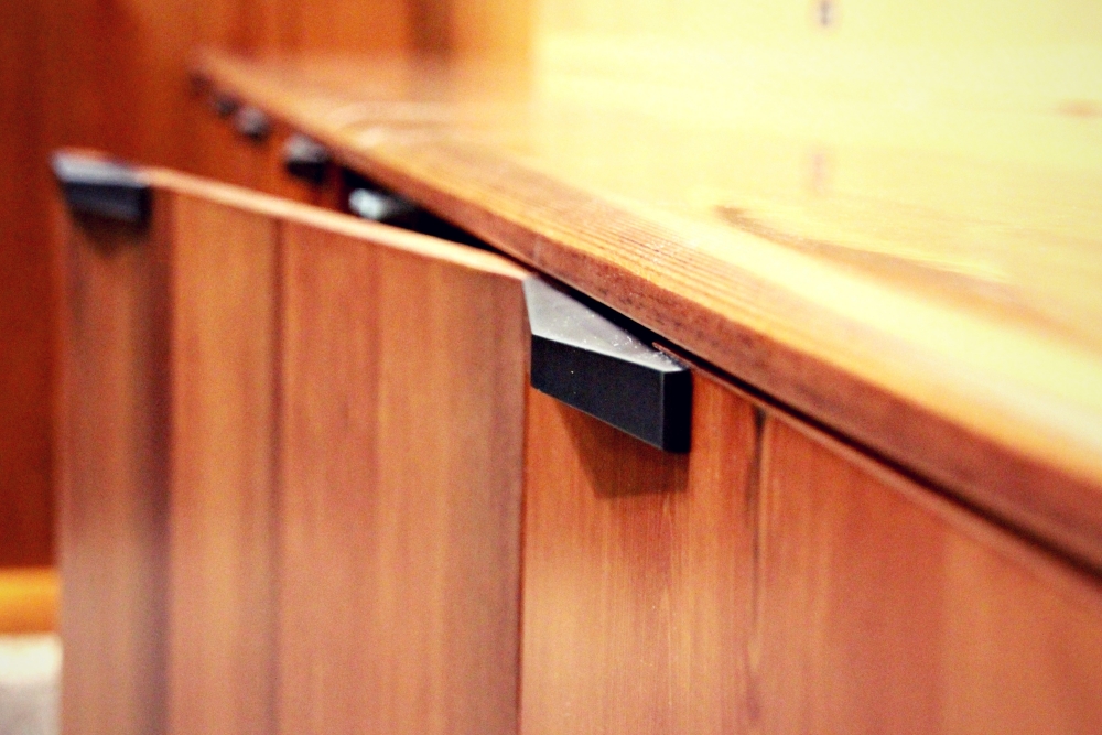 Close-up of a slightly ajar wooden cabinet door with black metal handle, featuring a glossy finish. The focus on the handle accentuates the clean lines and smooth texture of the cabinetry.