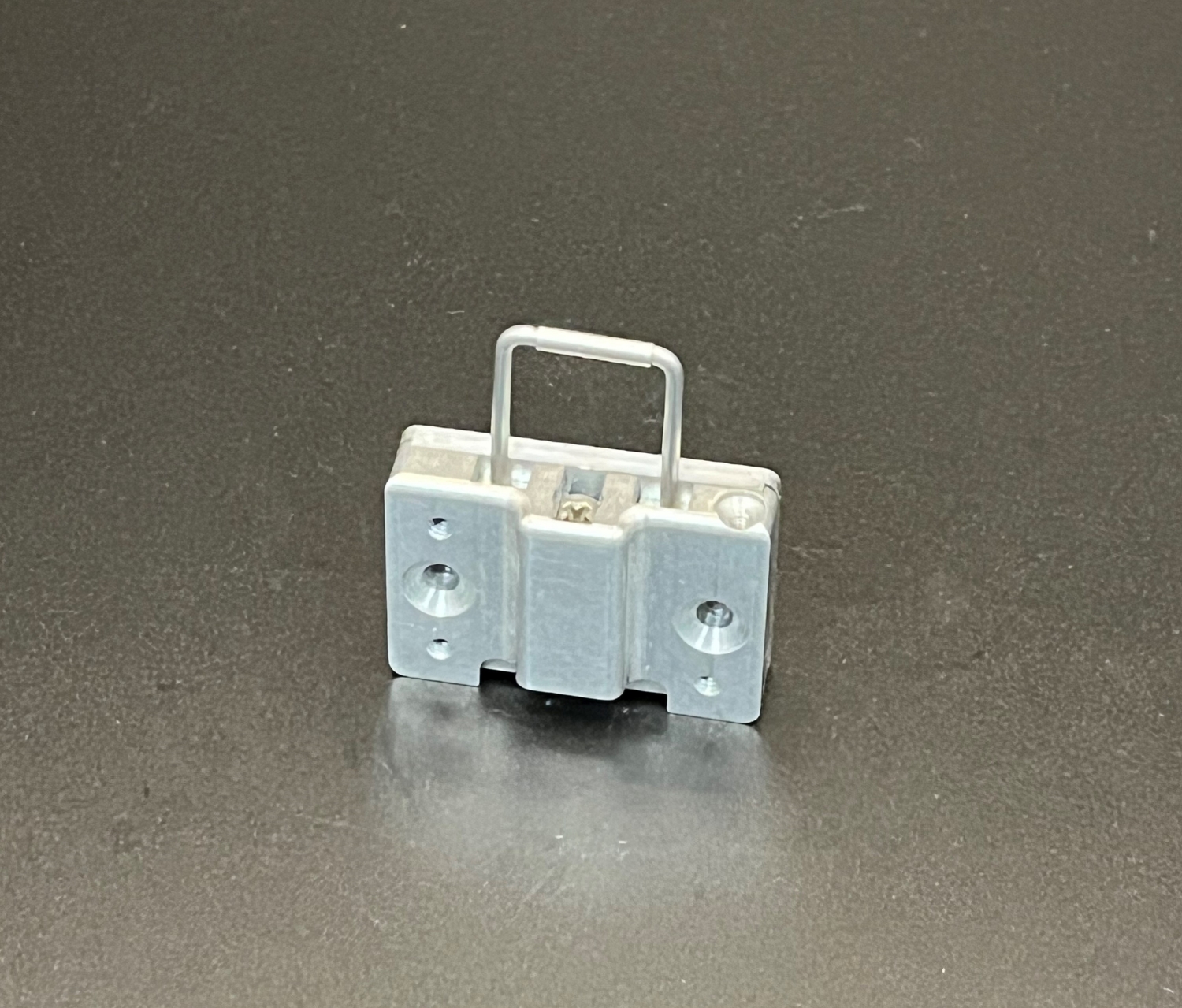 A small, rectangular metal component with two round holes and a U-shaped handle on a flat, dark surface.